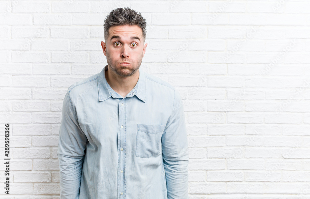 Young handsome man against a bricks wall blows cheeks, has tired expression. Facial expression concept. Young handsome man against a bricks wall blows cheeks, has tired expression. Facial expression concept.