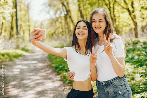 Two girls standing in a sunny forest. Friends have fun in a park. Ladies use the phone
