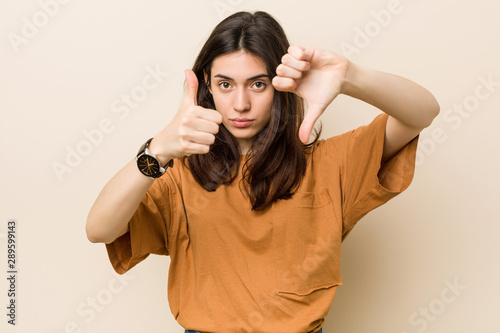Young brunette woman against a beige background showing thumbs up and thumbs down, difficult choose concept
