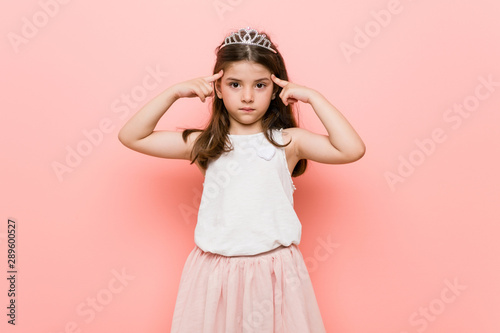 Little girl wearing a princess look focused on a task, keeping forefingers pointing head.