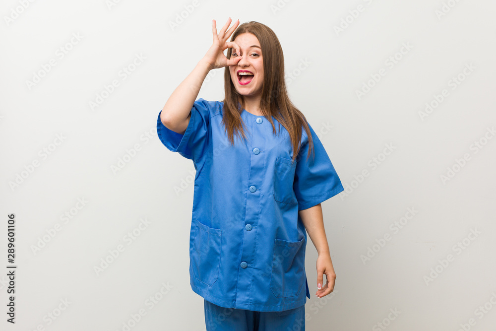 Young nurse woman against a white wall excited keeping ok gesture on eye. Young nurse woman against a white wall excited keeping ok gesture on eye.