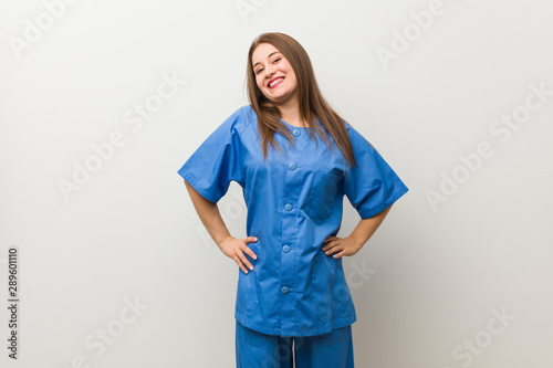 Young nurse woman against a white wall confident keeping hands on hips.