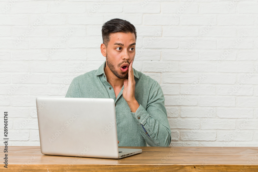 Young filipino man sitting working with his laptop is saying a secret hot braking news and looking aside Young filipino man sitting working with his laptop is saying a secret hot braking news and looking aside