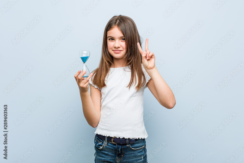Little caucasian girl holding an hourglass showing number two with fingers. Little caucasian girl holding an hourglass showing number two with fingers.