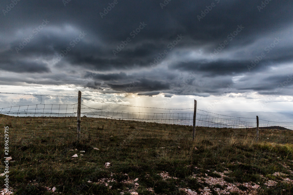 Sunray shines through clouds over the mountains in the middle of shadows, with a fence in the foreground Sunray shines through clouds over the mountains in the middle of shadows, with a fence in the foreground