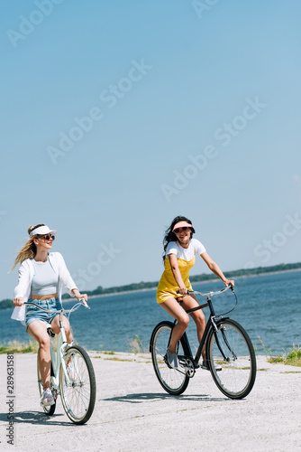 blonde and brunette girls riding bikes near river in summer
