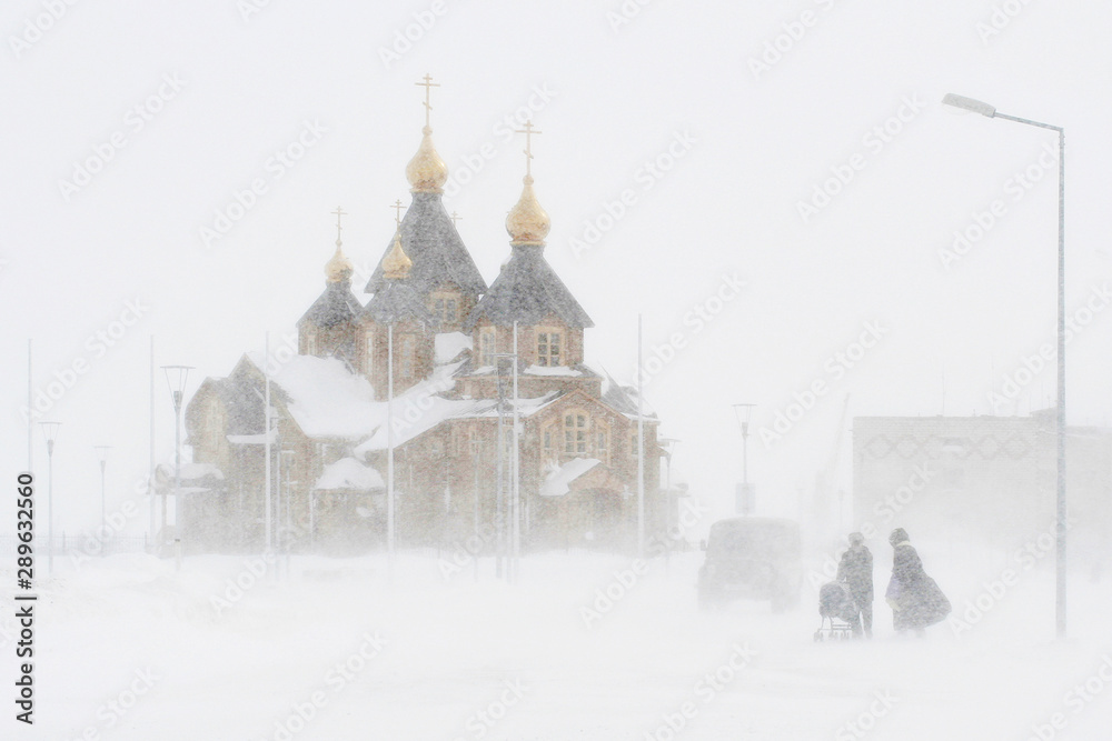 Snowstorm in the city. People during a blizzard are walking along the street. Strong wind and snowfall. Arctic climate. Cathedral of the Holy Trinity, Anadyr, Chukotka, Siberia, Far East Russia. April