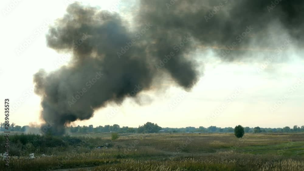 Column of black smoke in sky above field or meadow. This is fire, burns agricultural debris or explosion. Smoke swirls and is carried away by wind. Against of sunset and clouds. Outdoors.