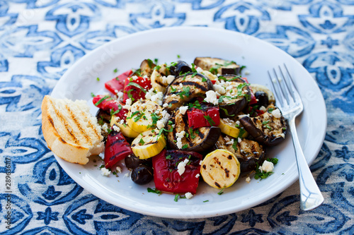 Grilled vegetables salad with feta cheese in white plate on blue textile background. Close up.
