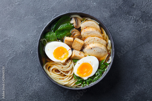 Asian noodle soup, ramen with chicken, tofu, vegetables and egg in black bowl. Top view. Copy space. Slate background.