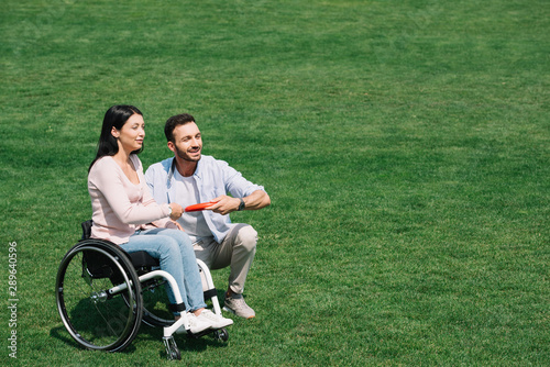 cheerful man holding flying disc while sitting near disabled girlfriend