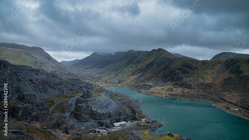Beautiful landscape image of Dinorwig Slate Mine and snowcapped Snowdon mountain in background during Winter in Snowdonia with Llyn Peris in foreground