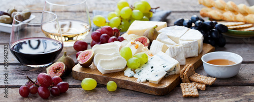 Cheese and fruits assortment on cutting board with red, white wine on wooden background.