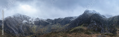 Stunning dramatic panoramic landscape image of snowcapped Glyders mountain range in Snowdonia during Winter with menacing low cloudshanging at the mountain peaks