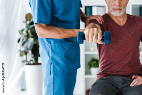 cropped view of doctor standing near middle aged man exercising with dumbbell