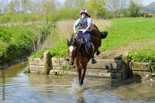 Young woman riding horse, jumping down a step into a water complex. 