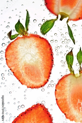 Fototapeta Naklejka Na Ścianę i Meble -  Sliced parts of a strawberry lie on a white background illuminated from above with water on it - background with fresh red strawberry with green on it in water with bubbles