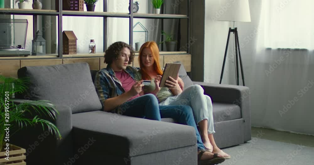 Young successful couple is busy with online shopping, using credit card and tablet computer, sitting on sofa at home, planning their purchase together, Caucasian. 4K, General plan, RED camera.