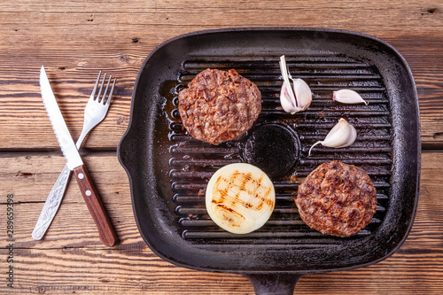 Fried burger beef cutlets with onion and garlic on grill pan on wooden background with fork and knife. Grilled food.