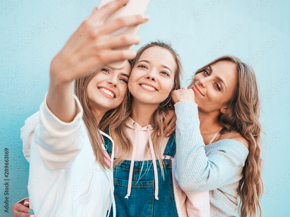 Three young smiling hipster women in summer clothes.Girls taking selfie self portrait photos on smartphone.Models posing in the street near wall.Female showing positive face emotions Three young smiling hipster women in summer clothes.Girls taking selfie self portrait photos on smartphone.Models posing in the street near wall.Female showing positive face emotions