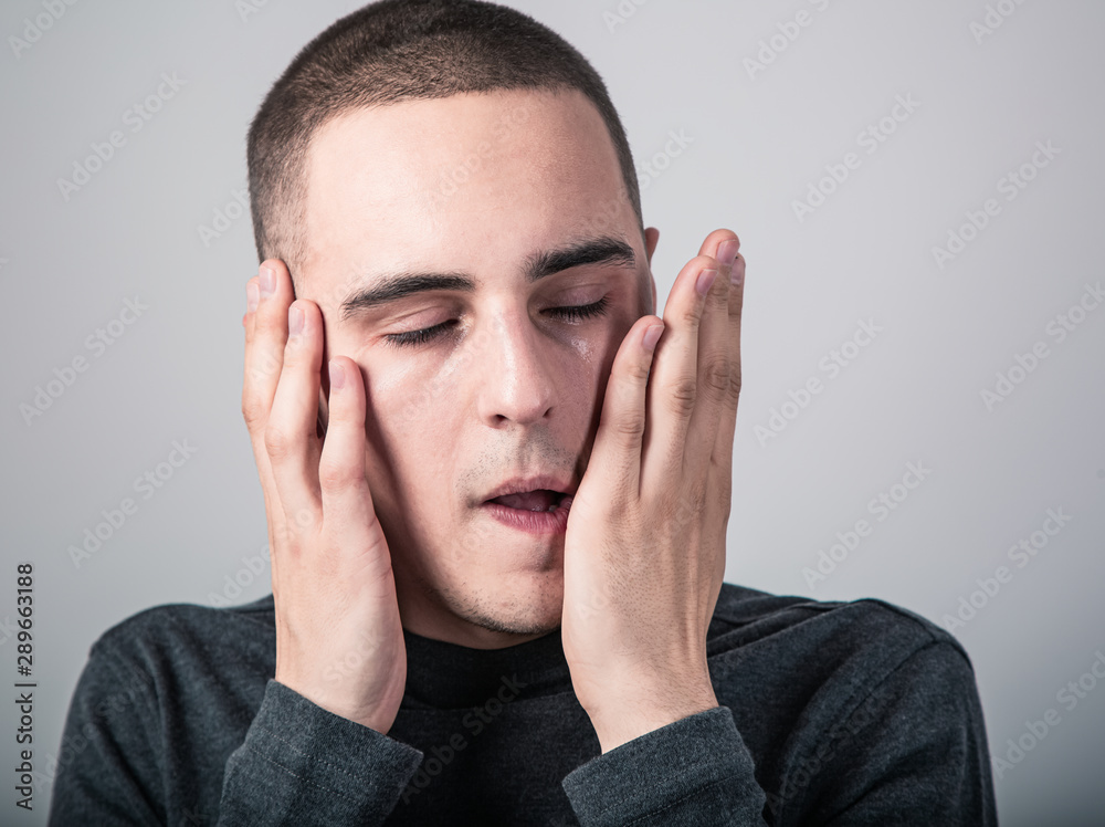 Unhappy crying man with dropping tears on the face touching the face the hand. Closeup studio portrait on grey background.