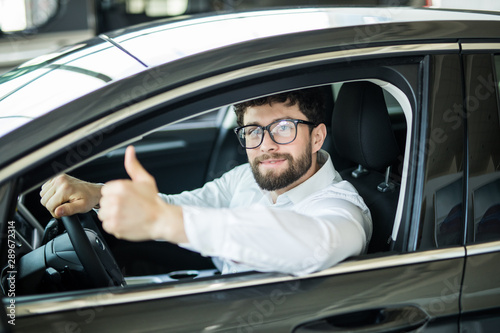 Happy handsome man smiling cheerfully showing thumbs up sitting in a new auto at the dealership salon copyspace car rental retail sales offer discount customer buyer consumerism.