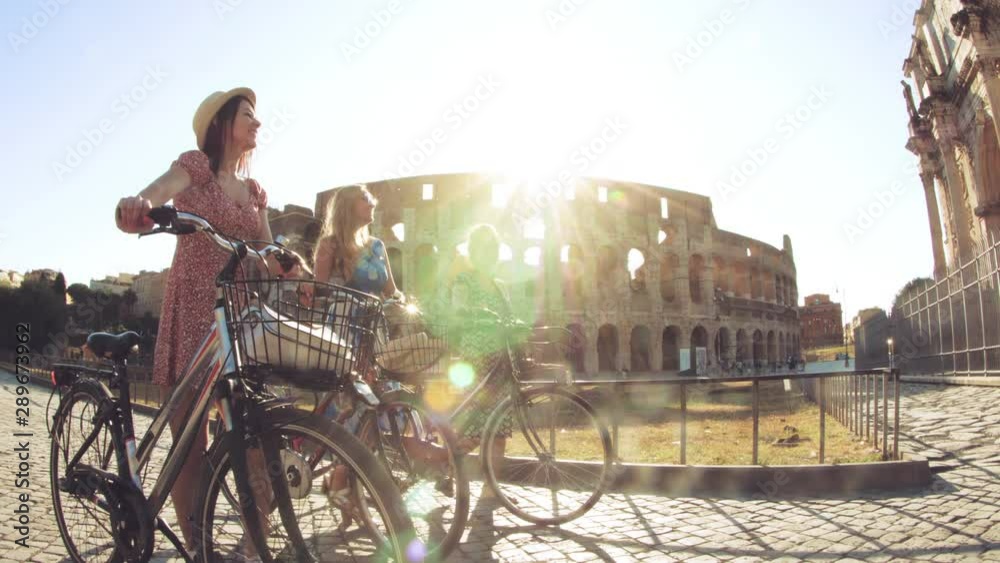 Three happy young women friends tourists walking with bikes at Colosseum in Rome, Italy at sunrise.