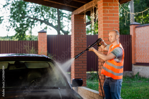 A working man in orange vest washes his car with a large head of water from a karcher on open air. High pressure cleaning
