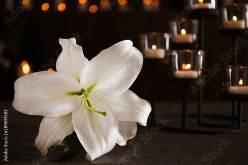 Fototapeta Naklejka Na Ścianę i Meble -  White lily and burning candles on table against blurred background. Funeral symbol