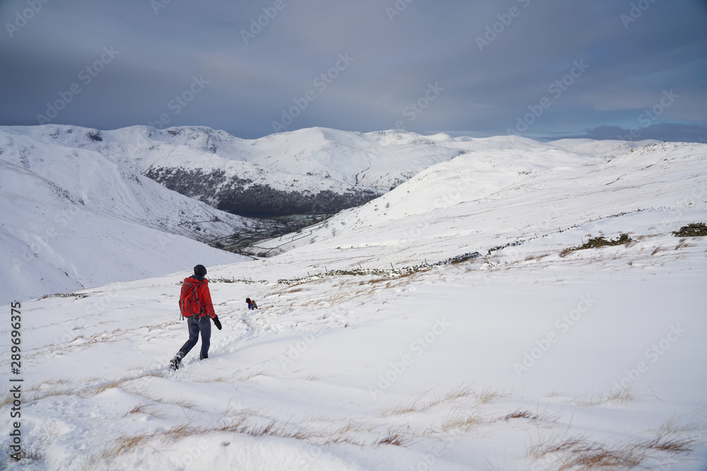 A hiker on the left edge of frame walking through fresh snow below the summit of The Knott near Hartsop in the Lake District.