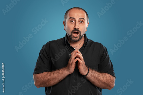 Close-up portrait of a brunet middle-aged man with beard, dressed in a black t-shirt and posing against a blue background.