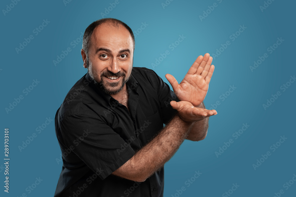 Close-up portrait of a brunet middle-aged man with beard, dressed in a black t-shirt and posing against a blue background. Close-up portrait of a brunet middle-aged man with beard, dressed in a black t-shirt and posing against a blue background.