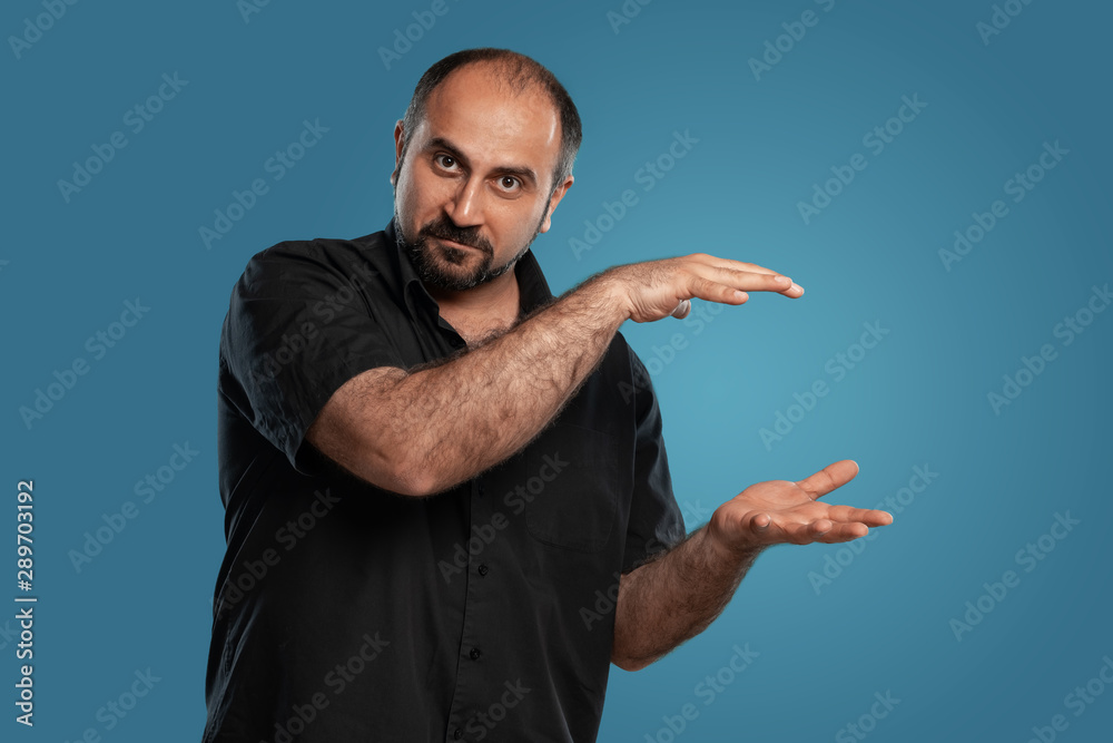 Close-up portrait of a brunet middle-aged man with beard, dressed in a black t-shirt and posing against a blue background. Close-up portrait of a brunet middle-aged man with beard, dressed in a black t-shirt and posing against a blue background.