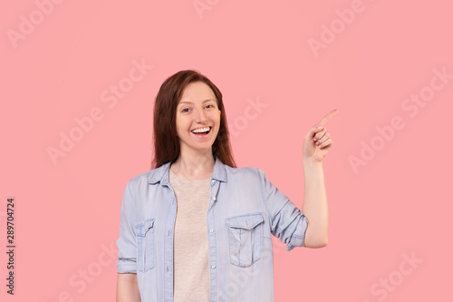 Smiling caucasian red-haired woman in full-day clothes posing on a pink background and pointing her finger at something useful. Young girl advertises goods and services. Place for text