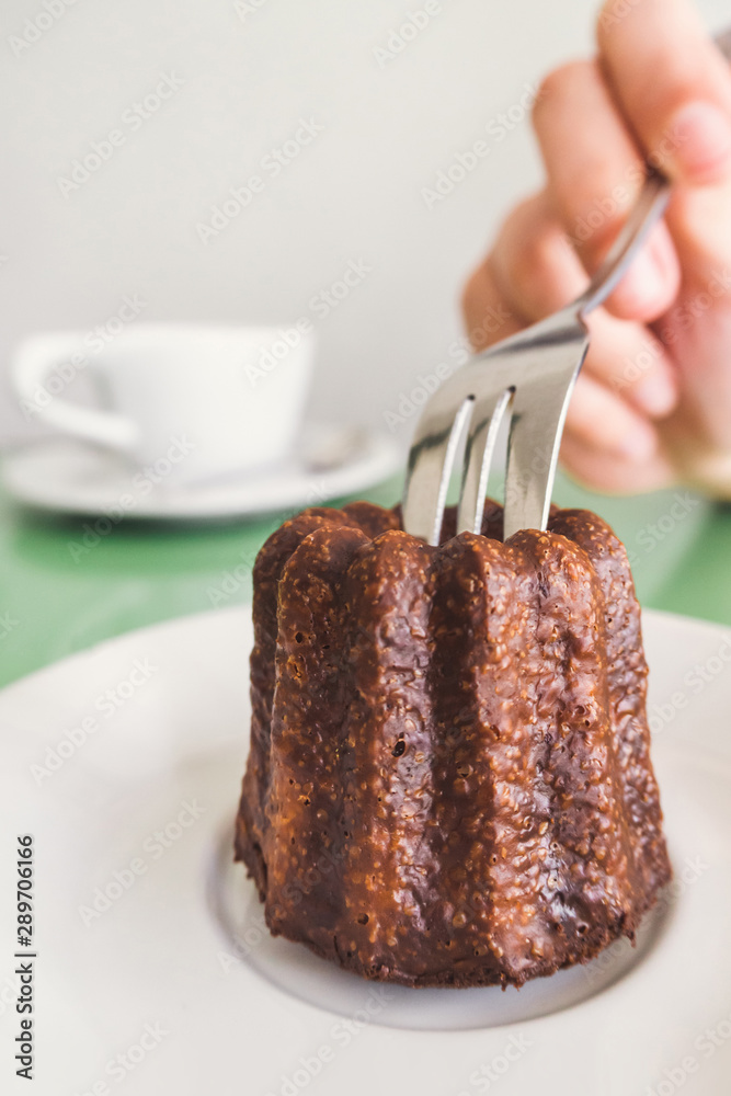 traveler woman hand holding a fork and cutting a Canelé (Canele) traditional French sweet dessert bakery with white coffee cup background in coffee shop in Paris. food and lifestyle concept. traveler woman hand holding a fork and cutting a Canelé (Canele) traditional French sweet dessert bakery with white coffee cup background in coffee shop in Paris. food and lifestyle concept.