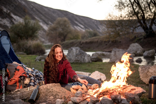 The young lady sits next to a fire in nature, does something to the fire and is very smiling