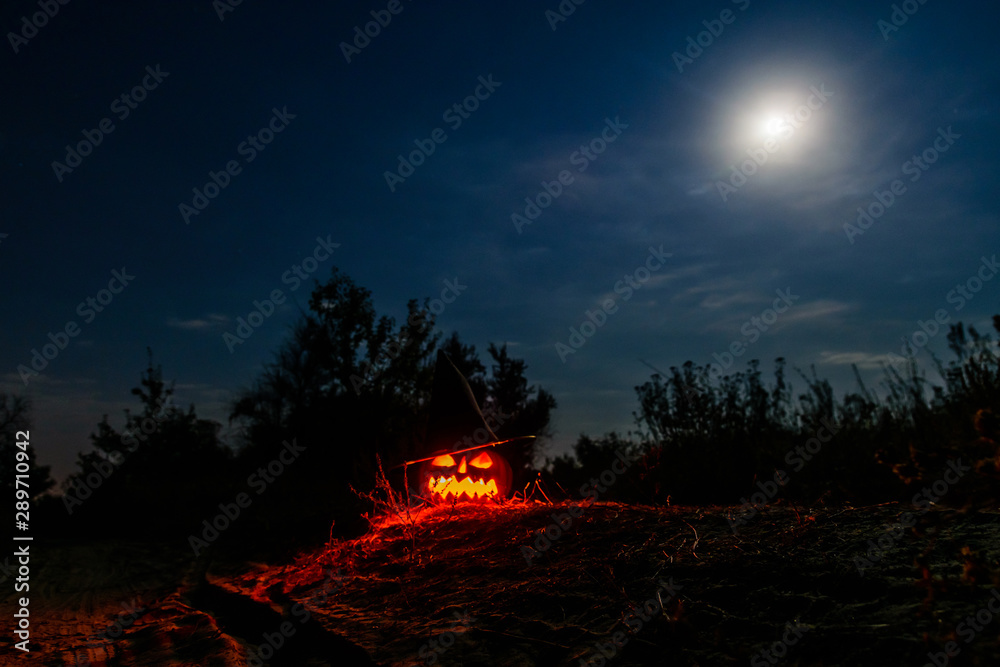 Fototapeta premium Carved halloween pumpkin jack-o-lantern wearing witch hat with burning candles glows in a darkness. Spooky landscape