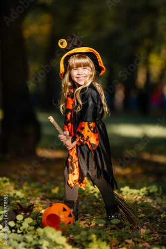 Little cheerful girl witch is standing in the park on the holiday of Halloween and holding a broom.