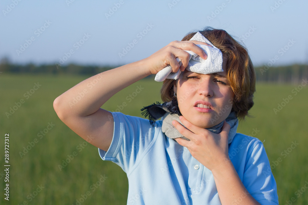 Naklejka premium Sick ill young woman in scarf is sweating suffering from a heat stroke. Girl has a sore throat outside in a summer park, in a hot warm day field. Female in fever, rubs her forehead with a handkerchief