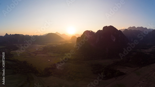 Aerial: Vang Vieng backpacker travel destination in Laos, Asia. Sunset over scenic cliffs and rock pinnacles, rice paddies valley, stunning landscape.