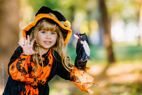 Halloween concept Cute little witch hiding behind a tree in a forest, park. Beautiful girl in witch costume with Halloween decoration.