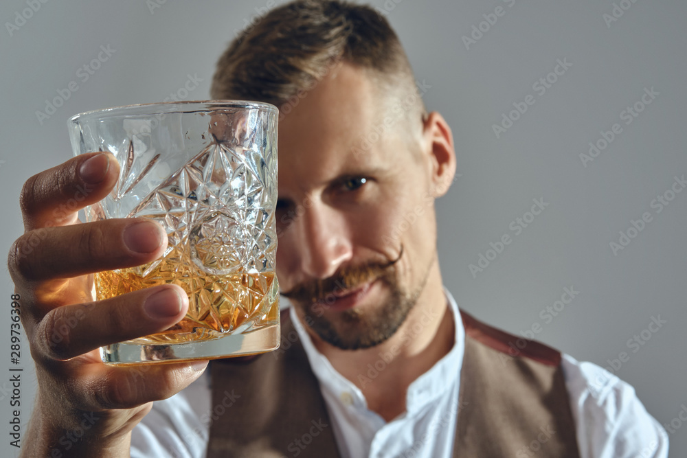 Man with stylish mustache, dressed in classic brown vest, white shirt is sitting at the table, enjoying whiskey. Grey background, close-up shot. Man with stylish mustache, dressed in classic brown vest, white shirt is sitting at the table, enjoying whiskey. Grey background, close-up shot.
