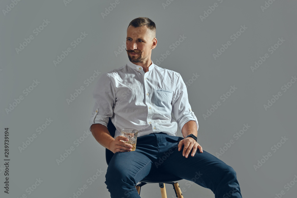 Man with stylish mustache, in classic white shirt and blue trousers is sitting on chair, holding glass of whiskey. Grey background, close-up shot. Man with stylish mustache, in classic white shirt and blue trousers is sitting on chair, holding glass of whiskey. Grey background, close-up shot.