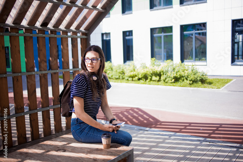 Young woman, a student, a brunette with glasses, sits on a park bench, takes a photo, looks around.