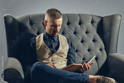 Man with stylish mustache, dressed in black shirt and trousers, beige vest is sitting on dark sofa, reading a magazine. Grey background, close-up.