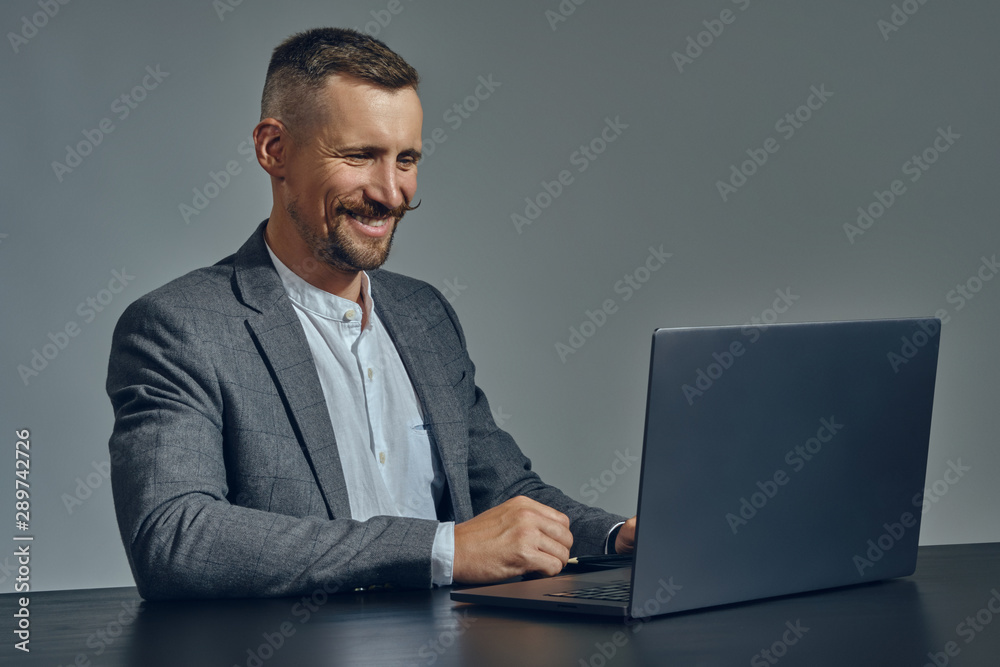 Bearded businessman with stylish mustache, dressed in a classic suit is working at laptop while sitting at table in office, gray background.