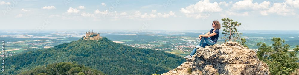 Landscape with young woman and Hohenzollern Castle, Germany. Adult girl travels in Alpine mountains in summer. Wide panorama of castle and person. - obrazy, fototapety, plakaty 
