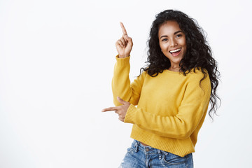 Cheerful carefree african-american woman in yellow sweater with curly haircut, smiling and laughing joyfully dancing as pointing sideways, showing left and up copy space, white background