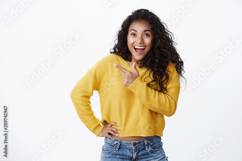 Inspired amused african-american woman yellow sweater gasping shocked and astonished, cheering emotionally telling great news, hold hand waist, pointing left blank space, standing white background photo