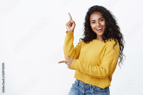 Cheerful carefree african-american woman in yellow sweater with curly haircut, smiling and laughing joyfully dancing as pointing sideways, showing left and up copy space, white background photo